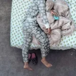 child wearing gray and white pajama lying on bed while holding phone