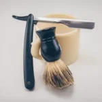 black straight razor beside beige ceramic mug and shaving cream brush