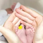 person holding baby's toe with yellow petaled flower in between