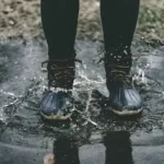 person wearing black duck boots stomping on body of water